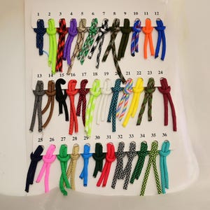 Peut inclure: Une carte de présentation présentant trente-six échantillons numérotés de paracorde dans diverses couleurs et motifs. Chaque corde est nouée et fixée sur la carte blanche, montrant des options incluant des couleurs unies, du camouflage et des motifs géométriques.