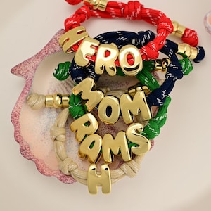 May include: A collection of four braided cord bracelets in red, navy blue, green, and beige. Each bracelet features gold-toned, rounded, metallic letter charms spelling out the words HERO, MOM, and RAMSH, secured with gold-toned metal end caps.