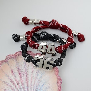 May include: Two adjustable cord bracelets featuring silver-toned metal number charms. One bracelet is dark red with white flecks and the number 44, while the other is black with white flecks and the number 36. Both have silver-toned metal end caps.