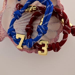 Pode incluir: Close-up de três pulseiras de estilo náutico. Uma é azul com detalhes brancos e o número 7 em dourado. Outra é bordô com detalhes dourados e o número 3 em dourado. A terceira pulseira é azul com detalhes brancos e detalhes dourados.
