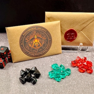 May include: A brown paper envelope with a red wax seal and a 5-card booster pack. The booster pack has a circular sticker with the text "Ashenborne Archive" and a logo. There are black dice with red pips, black gem-like pieces, and red and green gem-like pieces.