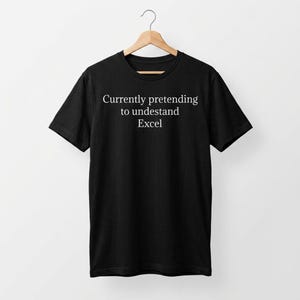 May include: A black short-sleeved t-shirt featuring the white text "Currently pretending to undestand Excel" printed on the chest. The garment is displayed on a wooden hanger, showcasing a crew neckline and a standard straight-cut silhouette.