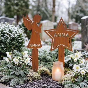 May include: Two rust-colored metal garden stakes featuring a cutout angel and a cutout star, both personalized with the name ANNA. The stakes are positioned upright in soil among green plants and a small frosted glass candle holder with a lit candle.