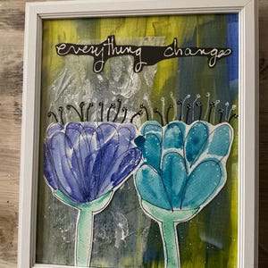 以下が含まれることがあります： 青とターコイズ色の水彩画の花が2つ描かれた額入りのアートワーク。背景は、黄色、青、緑の色合いの抽象的なデザインです。「everything changes」という言葉が上部に書かれています。