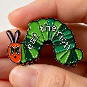 Könnte beinhalten: Ein farbenfroher Emaille-Pin in Form einer Zeichentrickraupe. Die Raupe ist grün mit Segmenten in verschiedenen Farbtönen, einem rot-orangen Kopf und den Worten "eat the rich" in Weiß.