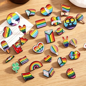 Könnte beinhalten: Eine Sammlung bunter Emaille-Anstecknadeln mit Regenbogen- und LGBTQ+-Pride-Themen. Die Anstecknadeln zeigen Herzen, Flaggen, Regenbogen und andere Symbole. Die Anstecknadeln liegen auf einer hellbraunen Holzoberfläche.
