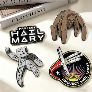 May include: Four enamel pins featuring designs from the book and movie, Project Hail Mary. Includes a black and white pin with the text "Project Hail Mary", a brown crab-like creature, a black and white figure, and a spaceship design.