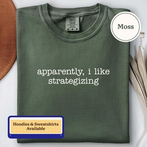 Può includere: Una t-shirt verde muschio con scollo rotondo e un'etichetta del marchio Comfort Colors all'interno del colletto. La parte anteriore della maglietta presenta il testo bianco "apparently, i like strategizing" in un semplice carattere sans-serif.