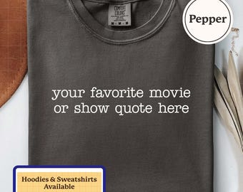 Puede incluir: Una camiseta de cuello redondo gris oscuro con el texto en blanco your favorite movie or show quote here impreso en el pecho. La prenda tiene una etiqueta de la marca Comfort Colors en el cuello y una etiqueta circular de color Pepper.