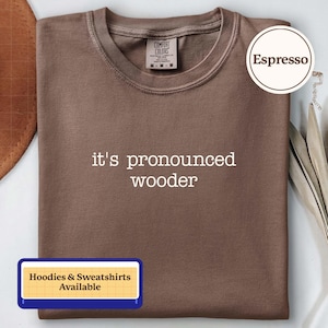 May include: An espresso-colored crew neck t-shirt featuring the white text 'it's pronounced wooder' printed on the chest. The shirt has a Comfort Colors brand label at the inner neckline indicating a size medium and 100% cotton material composition.