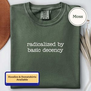 Puede incluir: Una camiseta de cuello redondo color verde musgo con el texto bordado en blanco "radicalized by basic decency" centrado en el pecho. La camiseta tiene una etiqueta de la marca Comfort Colors en el cuello interior que indica talla M y 100% algodón.