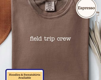 Puede incluir: Una camiseta de color espresso con el texto blanco 'field trip crew' impreso en el pecho. La prenda tiene cuello redondo y una etiqueta de la marca Comfort Colors en el interior del cuello. Una insignia circular dice 'Espresso' y un recuadro rectangular indica 'Hoodies & Sweatshirts Available'.