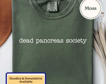 Puede incluir: Una camiseta de cuello redondo color verde musgo con el texto blanco dead pancreas society impreso en el pecho. La prenda incluye una etiqueta de la marca Comfort Colors en el cuello interior con la talla M. Una etiqueta circular muestra Moss.