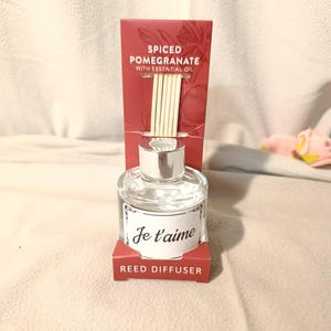 Op de afbeelding: Een heldere glazen rietverspreider met een zilveren dop en houten rietjes. Het label luidt "Je t'aime". De rode verpakking luidt "Spiced Pomegranate with Essential Oil" en "Reed Diffuser".