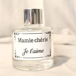 Peut inclure: Un flacon en verre transparent avec un bouchon argenté et une étiquette rectangulaire blanche. L'étiquette présente des coins décoratifs noirs et le texte imprimé en noir indique Mamie chérie Je t'aime en polices serif et script.