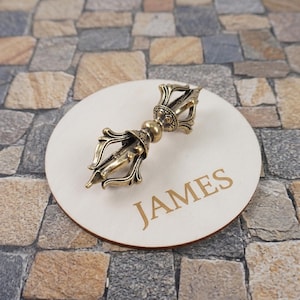 May include: A metallic gold-toned vajra ritual object with intricate symmetrical prongs resting on a circular light-coloured wooden disc. The wooden disc features the name JAMES engraved in a clear, capitalised serif font on its surface.
