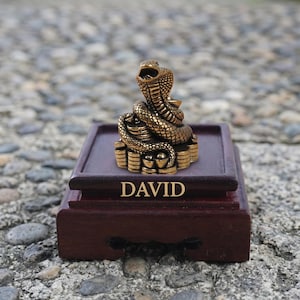 May include: A brass-coloured cobra figurine coiled atop a stack of coins, mounted on a dark brown wooden display base. The front of the wooden base features the name DAVID engraved in gold lettering, centred below the metallic snake sculpture.