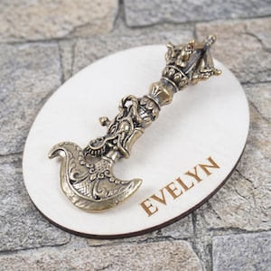 Puede incluir: Hacha decorativa de bronce con detalles de dragón, sobre un óvalo de madera clara. La palabra "EVELYN" está grabada en la madera. El hacha tiene una hoja curva y un mango ornamentado.
