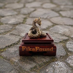 May include: A small, coiled brass snake figurine with detailed scales and a raised head, mounted on a dark reddish-brown wooden display base. The front of the wooden base features the text FikaFound engraved in a light-colored, sans-serif font.