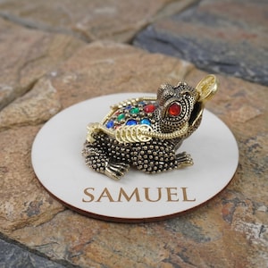 May include: A detailed, bronze-colored frog figurine with a coin in its mouth, adorned with colorful jewels. The frog sits on a white, circular wooden base with the name "SAMUEL" engraved in a brown font.