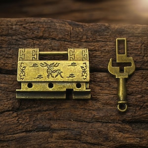 May include: An antique-style brass padlock featuring an engraved deer motif on the front plate, geometric border patterns, and a matching skeleton key with a rectangular top loop and a circular bottom ring, both displaying a weathered metallic finish.