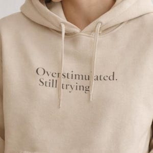 May include: A beige hoodie with a drawstring hood and a front pocket. The text "Overstimulated. Still trying" is printed in a dark brown color on the front of the hoodie.