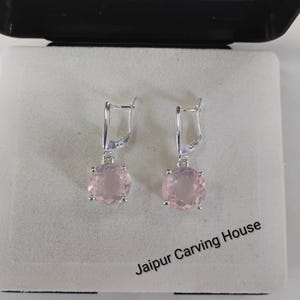May include: A pair of silver-toned drop earrings featuring round, faceted pale pink stones held by four-prong settings. The earrings are displayed in a white box with the text Jaipur Carving House printed in black on the lower right corner.