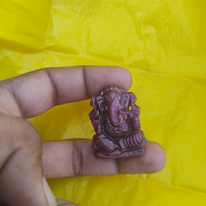 May include: A small, intricately carved figurine of the Hindu deity Ganesha, in a seated position. The figurine is a deep reddish-purple color and held against a bright yellow background.