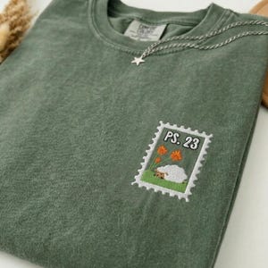 Puede incluir: Una camiseta de cuello redondo color verde salvia con un parche bordado en el pecho. El parche tiene forma de sello postal y muestra una oveja blanca sobre hierba verde con flores naranjas y el texto negro PS. 23 impreso arriba.