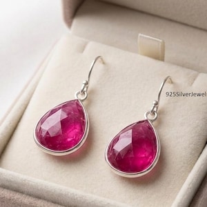 May include: A pair of silver earrings with teardrop-shaped, faceted, magenta-colored gemstones. The earrings are displayed in a beige jewelry box. The text "& SILVER EARRINGS" is visible at the top of the box. The seller's name, "925SilverJewelry26", is also visible.