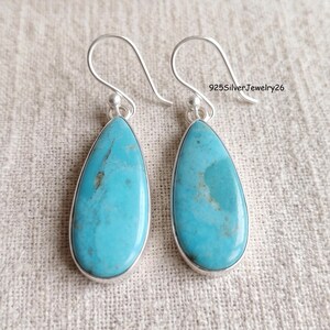 May include: A pair of teardrop-shaped turquoise earrings set in silver with curved ear wires. Each earring features a smooth, polished blue-green stone with natural brown matrix veining. The text 925SilverJewelry26 is visible in the upper right.