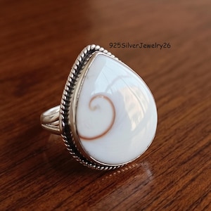 Puede incluir: Un anillo en forma de lágrima con una concha de ojo de Shiva blanca y lisa que presenta un patrón de espiral marrón natural. La piedra está engastada en un bisel de plata pulida con un borde de cuerda trenzada y banda dividida. Texto 925SilverJewelry26 visible.