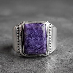 May include: A silver ring featuring a rectangular, polished purple charoite gemstone with swirling patterns. The stone is set in a silver bezel framed by beaded metal detailing on both sides. The text 925SilverJewelry26 is visible in the corner.