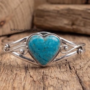 May include: A sterling silver cuff bracelet featuring a central heart-shaped turquoise stone with dark matrix veining. The silver band displays decorative swirling wire details on both sides of the stone. The text 925SilverJewelry26 is visible.