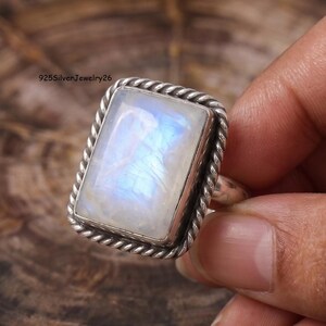May include: A rectangular ring featuring a large, iridescent moonstone cabochon set in a polished silver frame with a twisted rope-style border. The stone displays a soft blue glow against a white base. The text 925SilverJewelry26 is visible.