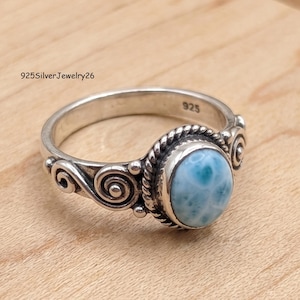May include: A sterling silver ring featuring an oval-shaped, light blue Larimar gemstone set in a twisted rope border. The band is decorated with silver scrollwork and spiral patterns on both sides. The interior of the band is stamped with 925.
