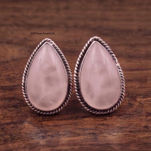 Könnte beinhalten: Ein Paar tropfenförmige Ohrstecker mit glatten, blassrosa Rosenquarzsteinen in poliertem Sterlingsilber. Jeder Stein ist von einem dekorativen gedrehten Seilrand umgeben, und der Text 925SilverJewelry26 ist deutlich zu erkennen.
