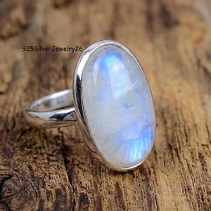 May include: A sterling silver ring featuring a large, oval-shaped moonstone cabochon with a translucent white appearance and blue adularescence. The metal band is smooth and polished. The text 925SilverJewelry26 is visible in the upper left corner.
