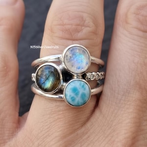 May include: A sterling silver ring set featuring three round cabochon gemstones: a translucent moonstone, a dark labradorite with blue iridescence, and a light blue larimar. The band includes the text 925SilverJewelry26 and a textured detail.