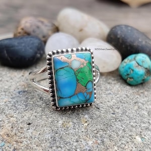 May include: A sterling silver ring featuring a rectangular, multi-colored turquoise stone with blue and green hues and copper-colored matrix veins. The stone is set in a beaded silver frame with a split band design. Text 925SilverJewelry26 is visible.