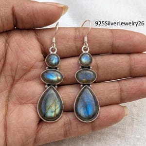 May include: Silver earrings featuring three labradorite stones. The top stone is round, the middle is oval, and the bottom is a teardrop shape. The stones display blue and gold iridescence. The earrings are on a person's hand.