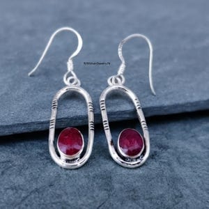 May include: A pair of silver drop earrings featuring an elongated oval frame with etched horizontal line details. Each earring holds a single, faceted, deep red oval gemstone at the base. The text 925SilverJewelry26 is visible between the hooks.