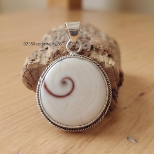 Puede incluir: Un colgante redondo con una concha de ojo de Shiva blanca con un patrón de espiral marrón natural, engastado en un marco de plata trenzado con una anilla de plata en la parte superior. El texto 925SilverJewelry26 es visible arriba a la izquierda.