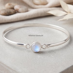 May include: A sterling silver bangle bracelet featuring a central round moonstone cabochon set in a smooth bezel. The gemstone displays a soft blue adularescence. The text 925SilverJewelry26 is visible above the stone on the silver band.