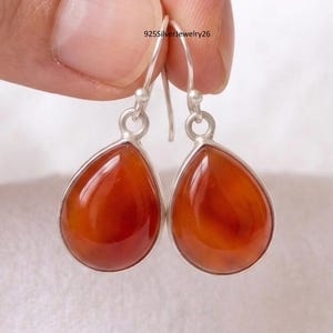May include: A pair of silver earrings with teardrop-shaped, orange-red gemstones. The gemstones are set in a silver bezel and have a polished, glossy finish. The earrings are suspended from silver ear wires. The text "925SilverJewelry26" is visible in the background.