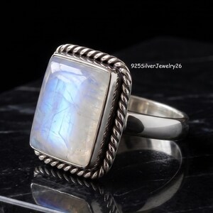 May include: A sterling silver ring featuring a large, rectangular, iridescent moonstone cabochon set within a twisted rope border. The band is smooth and polished. The text 925SilverJewelry26 is visible in the upper right corner of the image.