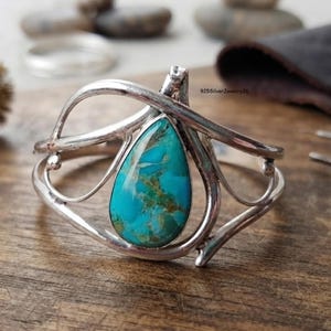 May include: A silver cuff bracelet featuring a central teardrop-shaped turquoise stone with brown matrix veining. The metal band has a polished finish and an open, swirling wire design that frames the stone. The text 925SilverJewelry26 is visible.