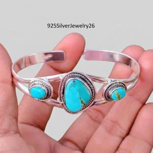 May include: A silver cuff bracelet featuring three turquoise stones. The central stone is oval, flanked by two smaller, round stones. The bracelet is held in a hand against a light background. The image includes the text "925SilverJewelry26".