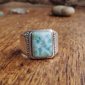 May include: A silver ring featuring a rectangular, light blue and white marbled stone set in the center. The metal band has a beaded texture along the sides of the stone setting. The text 925silverJewelry26 is visible near the ring.