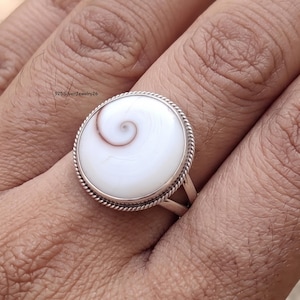 May include: A silver ring featuring a round, white Shiva eye shell with a natural brown spiral pattern. The shell is set in a polished silver bezel with a decorative twisted rope border. The band is made of two thin, smooth silver wires.